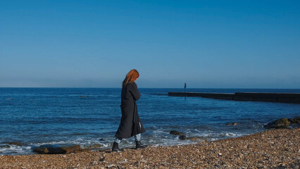 Person walking along the beach shore on a sunny day with calm waves and another figure in the distance near a pier