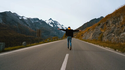 A person walks confidently down a winding road surrounded by mountains and snow, capturing a sense...