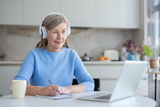 Senior woman with headphones studies online from her bright home kitchen, smiling and focused while taking notes on a laptop during a webinar or virtual class, lifelong learning