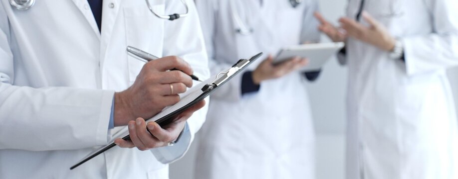 Doctor writing on a clipboard during a medical team discussion, collaborating on patient care, documentation, and healthcare strategy in a clinical setting - Powered by Adobe
