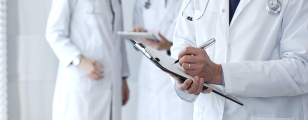 Doctor taking notes on clipboard during discussion with medical team involved in collaborative patient care, documentation, and health care strategy development in clinic