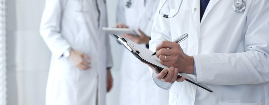 Doctor taking notes on clipboard during discussion with medical team involved in collaborative patient care, documentation, and health care strategy development in clinic