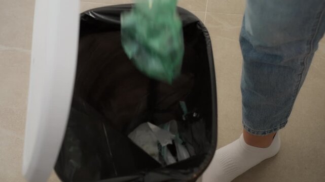 Person open white pedal trash can with their foot and throws green crushed plastic bottle inside, promoting recycling and proper waste disposal in modern household setting
