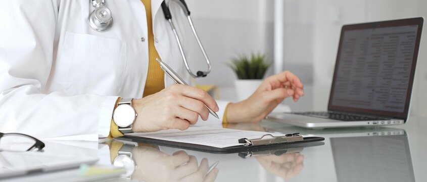 Doctor working on medical records, writing prescription in clinic office, using laptop for patient data management. Medicine, healthcare and science concept - Powered by Adobe
