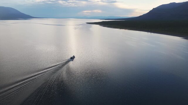 Drone tracks a lone vessel over silver water at dusk on Lake Lama. The wake draws leading lines to the horizon while the Putorana Plateau frames the route through pristine Siberian wilderness