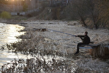 Boy fishing from a small wooden pier on a calm lake at sunset, surrounded by dry reeds and golden reflections on the water — peaceful spring or autumn evening outdoor scene