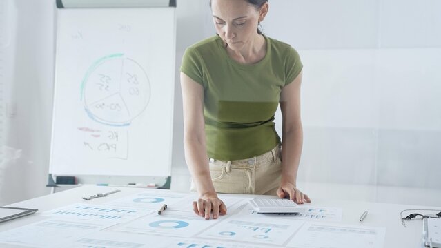 Businesswoman pointing at financial charts and graphs on a table, with a pie chart on a flip chart in the background. Business people concept