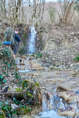 A hiker with a large backpack contemplates a small waterfall in early spring's bare forest, capturing nature's quiet power.