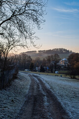 Obraz premium Rural dirt road covered with frost leading toward village and hills on a calm winter sunrise morning.