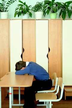 Schoolboy with head down on desk in classroom, surrounded by wooden lockers and plants, expressing feelings of sadness and isolation due to bullying at school