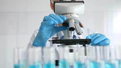 Female scientist wearing protective equipment checking microscope, analyzing blue test tubes in modern laboratory research setting. Medicine, healthcare and science concept