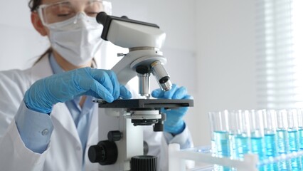 Female scientist wearing protective equipment checking microscope, analyzing blue test tubes in modern laboratory research setting. Medicine, healthcare and science concept