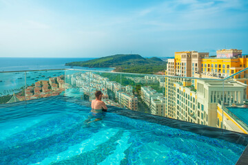 Man relaxing in rooftop infinity pool with stunning panoramic view of tropical island and Mediterranean-style buildings in Sunset town on Phu Quoc, Vietnam. Vacation, relaxation, tropical resort