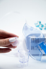 Vertical photo, close-up of a medicinal solution dripping from an ampoule into an inhalation bowl against the background of a blue breathing mask and a device on a white background. The concept of