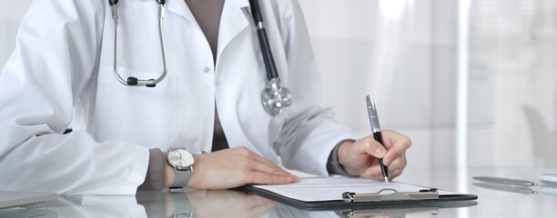 Female doctor in white coat with stethoscope writing patient notes on clipboard, completing medical records in clinic office. Medicine and health care