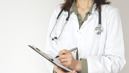 Woman doctor, wearing a stethoscope and a medical coat over a green blouse, is standing and writing...