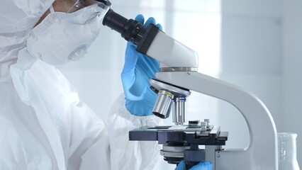 Medical virologist in a hazmat suit, mask, and gloves, diligently working at a microscope in a laboratory researching viruses. Healthcare and science concept