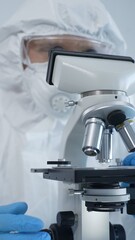Male scientist virologist wearing hazmat suit, mask, and blue gloves, diligently working at a microscope in a laboratory researching viruses. Medicine and science concept
