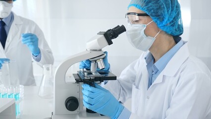 Woman scientist in blue protective gear is examining specimen under microscope while laboratory...