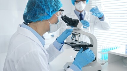Woman scientist in blue protective gear is examining specimen under microscope while laboratory assistant handles glassware in medical investigation at the background. Medicine, and science concept