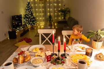 Thoughtful young boy at festive Christmas dinner table with lit red candles, holiday treats and decorated tree with fairy lights in warm cozy home interior. Family celebration concept.