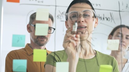Professional team brainstorming project strategy, placing colorful sticky notes on glass board, analyzing data, sharing insights, developing collaborative solutions