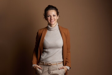 A smiling businesswoman in her 30s-40s, dressed in a brown blazer and beige trousers, stands with...