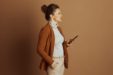 A sophisticated businesswoman in a brown blazer and cream turtleneck stands in profile, thoughtfully engaged with her black smartphone. Her pose conveys modern connectivity and a quiet elegance.