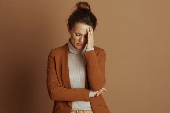 A professional woman, in a brown blazer and beige turtleneck, looks down with a pained expression and hand to her forehead, conveying discomfort, stress, or exhaustion against a brown background.