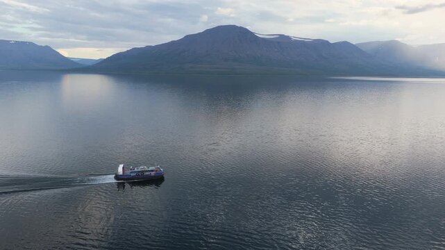 Aerial view of a hovercraft running on calm Lake Lama as a volcanic table mountain looms across the bay in soft evening light. Slow pull reveals scale, mirror water and the remote Siberian mood of the