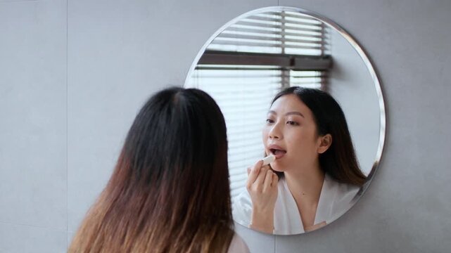 A young Asian woman applies skincare products in her bathroom, focused on her reflection in the mirror. She practices self care and relaxation in this serene environment.