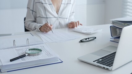 Female accountant is working with financial documents, using calculator and taking notes, with a magnifying glass and a folder on a white table in a bright office. Audit and taxes in business
