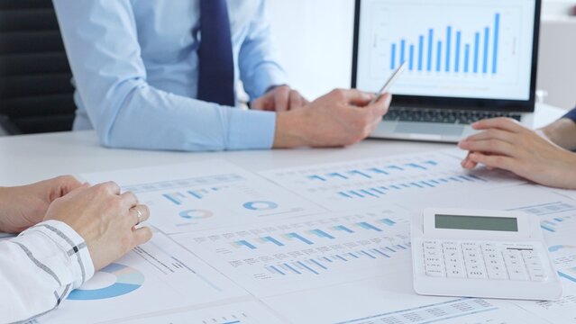 Businessman professional pointing laptop screen showing bar graphs, calculator on table while team collaborates on financial analysis and planning. Business people concept