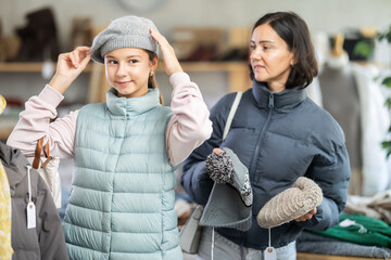 Adolescence girl try on hat in front of mirror during shopping with mother in clothes store. Customer is trying on clothes in store. Buyer is immersed in choice of clothes.