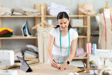Adult woman dressmaker draws pattern on paper according to drawing on tablet in workshop
