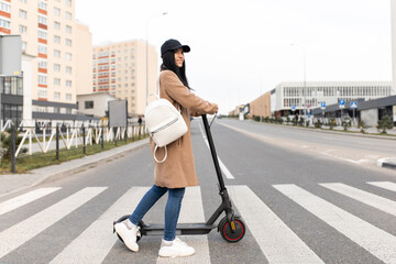A young woman in a beige coat walks with an electric scooter through a pedestrian crossing in the city. Eco-friendly transport. © zadorozhna