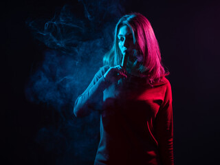 Obraz premium Woman vaper in dark room. Woman inhales vapor from mouth. Young girl smokes vape. Student with vape device in puff of smoke. Concept smoking e-cigarette instead tobacco. Woman vape illuminates light