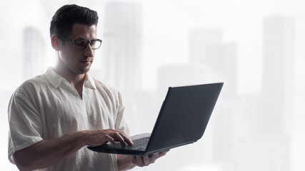 Business man holding laptop. Manager standing using computer. Young guy in shirt near blurred skyscrapers. Programmer from IT company. System administrator with laptop. Business man using internet