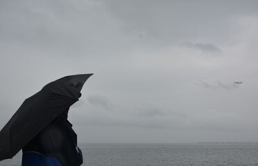 A woman stands under an umbrella near the sea in cloudy weather and looks at a plane flying away into the distance