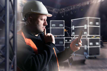 Stage supervisor in helmet and safety vest uses smartphone to coordinate equipment setup, managing...