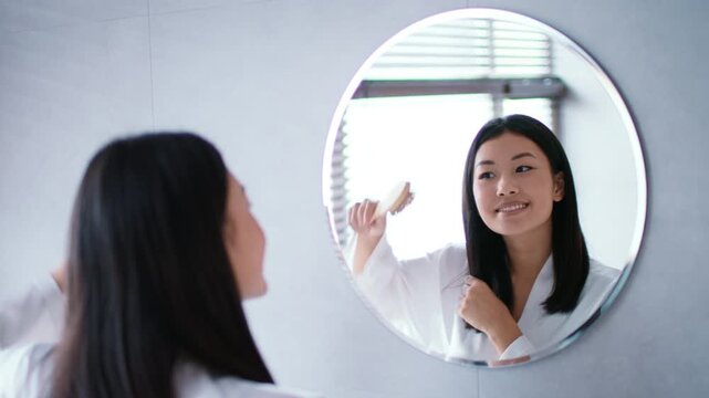 In a bright bathroom, a young Asian woman is pampering herself, smiling as she applies hair care products while admiring her reflection in the round mirror, combing her hair