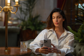 Young businesswoman using smartphone sitting on cushioned chair in hotel lobby. Woman in white shirt and appears relaxed. Warm lighting create cozy atmosphere