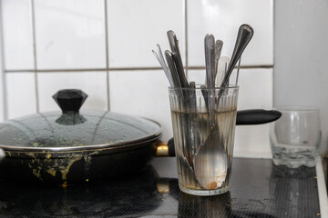 Used kitchen utensils soak in a glass of water next to a soiled frying pan on a dark stovetop surface.