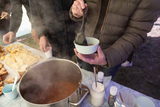 A person serves steaming hot food from a large pot into a small bowl on an outdoor table, surrounded by other snacks.