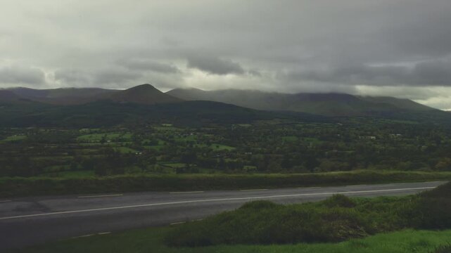 OA dramatic and moody landscape captures a panoramic view of rolling hills and mountains under a heavy, overcast sky. The dark, ominous clouds hang low, partially obscuring the mountain peaks and