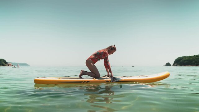 Woman in swimsuit for surfing stands up and learns to paddle board on turquoise sea, balancing swim. Concept captures the essence of summer adventure and the joy of trying something new.