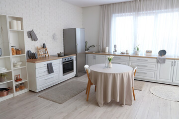 Interior of light kitchen with  table, white counters and silver fridge near window