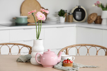 Pink teapot, tasty cake and vase with flowers on table in kitchen