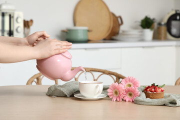 Young woman pouring hot tea from pink teapot into cup in kitchen
