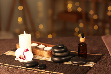 Bamboo mat with spa stones, candles, cosmetic product and orchid flower on table against blurred garland in dark salon
